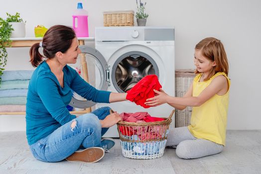 Mother with daughter sitting on floor near washing machine with clothes in basket in light bathroom