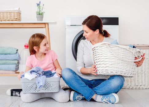 Little girl helping mother with heap of clothes during washing holding basket with laundry