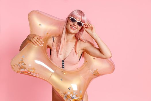 Happy young woman with pink hair in swimsuit and sunglasses with inflatable star for swimming posing isolated over pink background. Summer vacation, fun concept