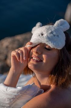 Young woman in sleep mask posing on the seashore. She is wrapped in a white blanket. The coast of the sea at a height, around the sea and rocks, dawn