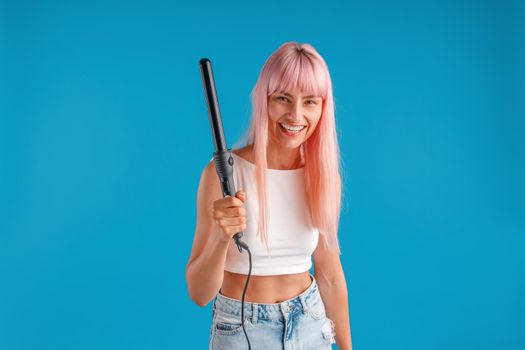 Joyful young woman with pink hair in casual wear smiling at camera, holding curling iron, standing isolated over blue studio background. Beauty and hair care concept