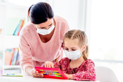 Teacher and schoolgirl in protective masks counting on abacus in light classroom