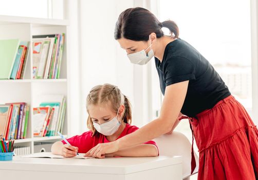 Little girl writing in notebook with teacher's help at school during virus epidemic