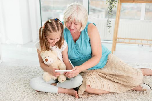 Happy grandmother with little granddaughter using tablet siting on floor in children's room