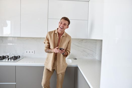 Hadsome smiling man is chilling on the minimalistic kitchen with smartphone