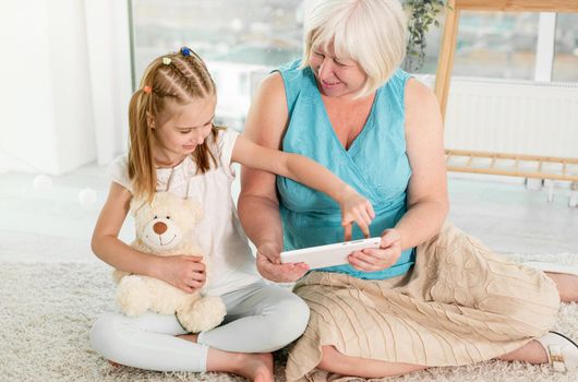 Cute little girl teaching granny to use tablet sitting on floor in light room