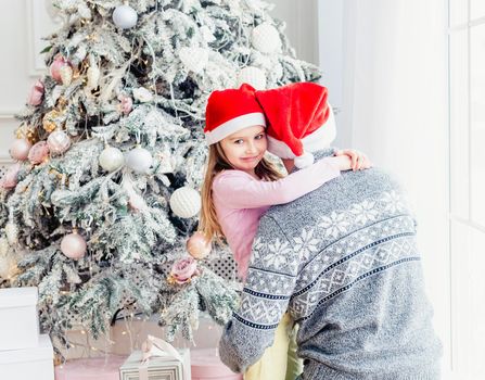 Little daughter hugging father in santa hat near christmas tree