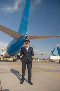 Full length shot of professional pilot in uniform looking at camera, standing in front of big passenger airplane ready for departure in airport. Aircraft, occupation, transportation concept