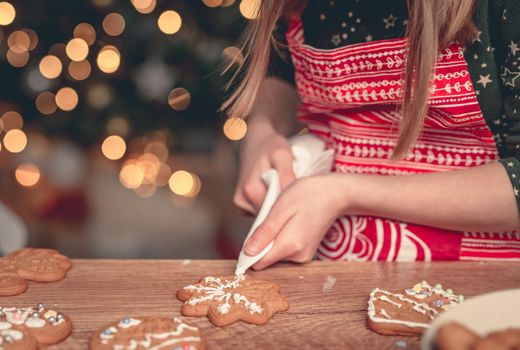 Girl in festive apron decorating gingerbread snowflake using culinary bag at home