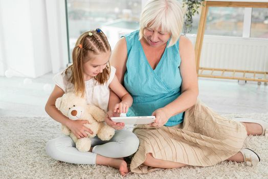 Happy grandmother with little granddaughter using tablet siting on floor in children's room