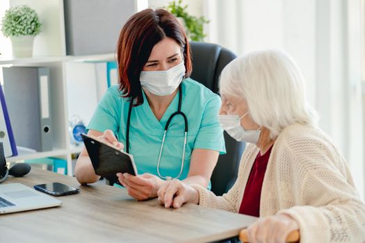 Doctor showing prescriptions on paper to aged woman patient in protective mask during appointment in clinic