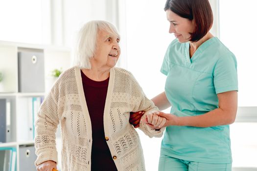 Smiling caregiver helping happy elderly woman to walk in nursing home