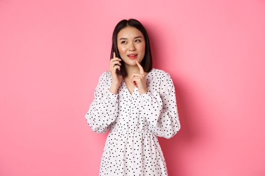 Thoughtful asian woman calling on mobile phone to make order, thinking and looking aside, standing against pink background.