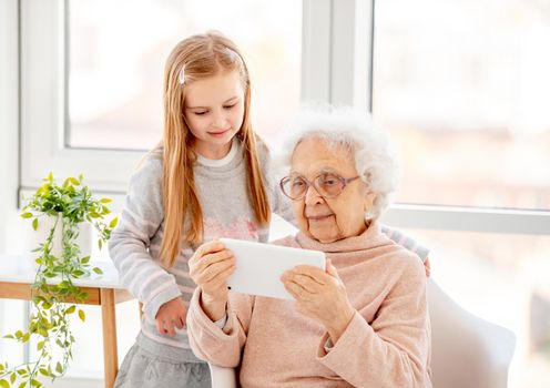 Cute school girl helping old lady with gadget in light room