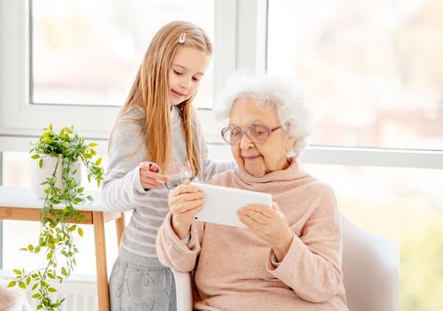 Cute school girl helping old lady with gadget in light room