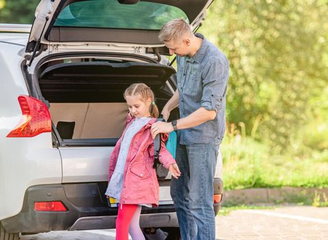 Father helping take off daughter's backpack after school near car trunk