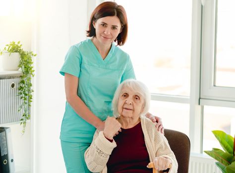 Caregiver standing beside elderly woman on chair in light room inside nursing home