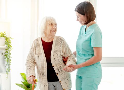 Smiling caregiver helping happy elderly woman to walk in nursing home