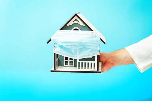 Model house in the hands of a doctor on blue background, house with protective medical mask. Safety to stay at home