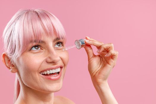 Close up portrait of cute woman with pink hair smiling while applying serum or hyaluronic acid on her skin using dropper isolated over pink background. Beauty, cosmetics and skincare concept