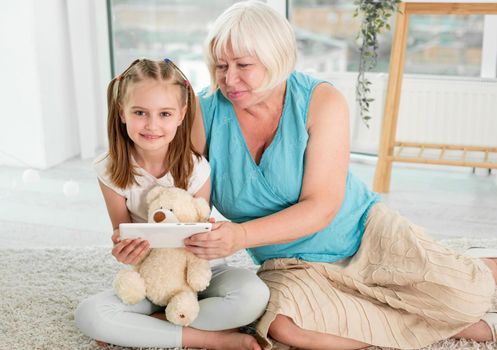 Happy grandmother with little granddaughter using tablet siting on floor in children's room