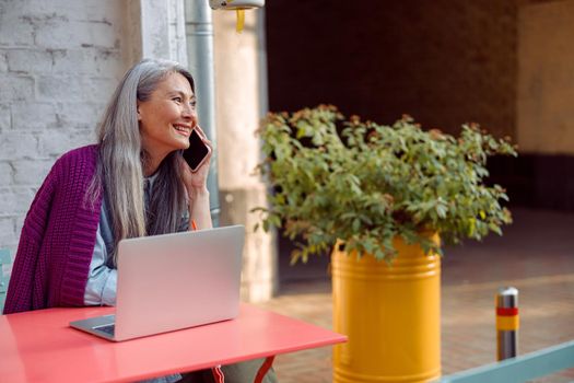 Happy senior Asian woman with long hair talks on mobile phone sitting at table with laptop on outdoors cafe terrace