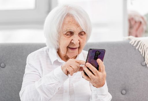 Elderly woman sitting on the sofa at home with smartphone and touching screen with her finger