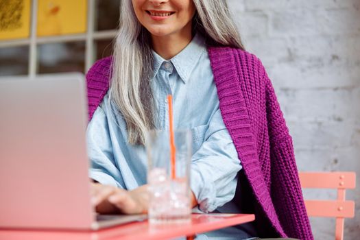Smiling mature woman with grey hair works on modern laptop sitting at color table on outdoors cafe terrace closeup