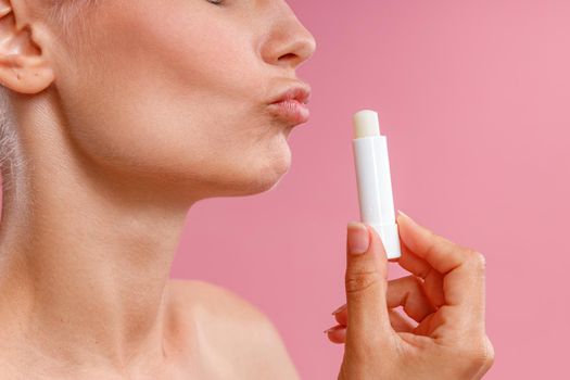 Close up shot of woman holding lip balm near her lips and making kissing gesture, posing isolated over pink background. Beauty, lip care concept. Side view