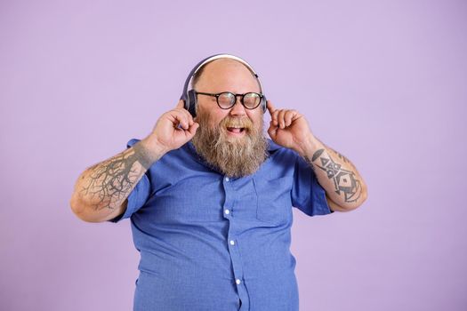 Happy bearded man with overweight in tight shirt listens to music with modern headphones posing on purple background in studio