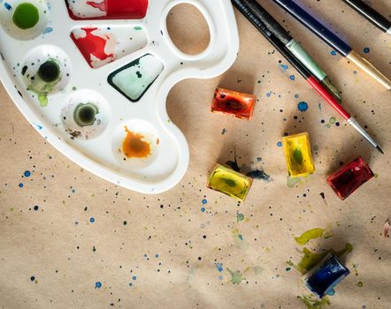 Plastic art palette with used brushes and opened paint boxes on paper-covered workshop table