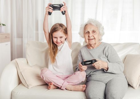 Smiling grandmother with little granddaughter playing games together with gamepads