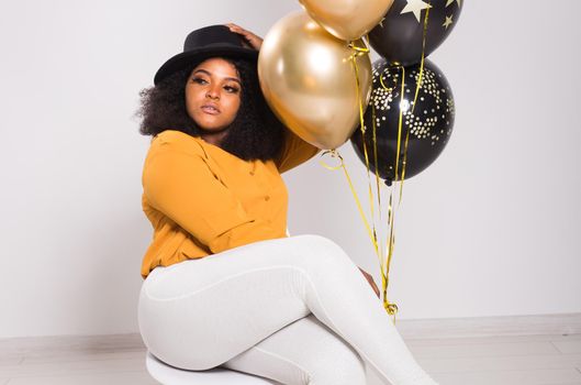 Portrait of smiling young African-American adult woman looking sweet on yellow background holding balloons.