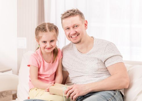 Smiling dad with pretty daughter opening cute Fathers day gift on classy sofa