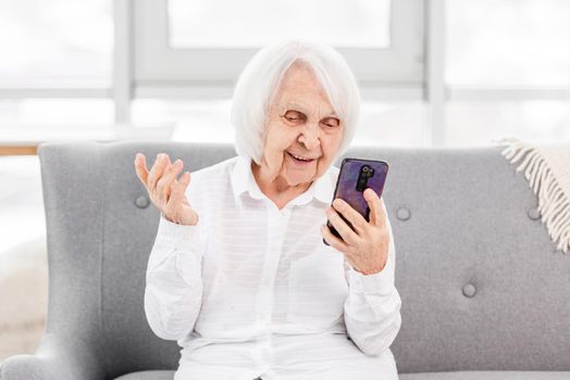 Modern ederly woman sitting at the sofa at home and talking with friends by video using smartphone