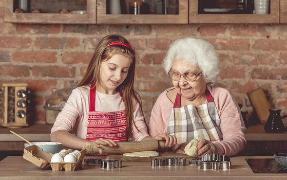 Elderly woman teaching a little girl in red apron to bake homemade cookies spreading dough with rolling pin