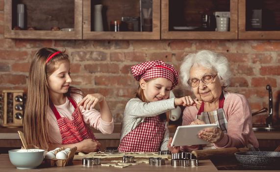 Grandma with her little granddaughters looking for a recipe using modern tablet