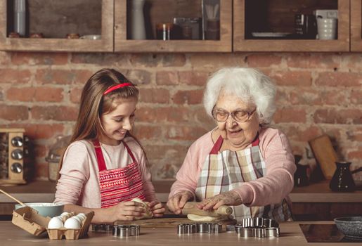 Elderly woman teaching a little girl in red apron to bake homemade cookies spreading dough with rolling pin