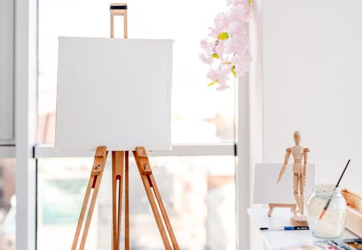 Wooden easel stands in the sunny white room workplace with painting tools on background