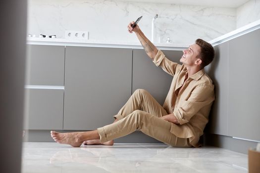 Beautiful caucasian man is sitting on the floor and using smartphone on light modern kitchen