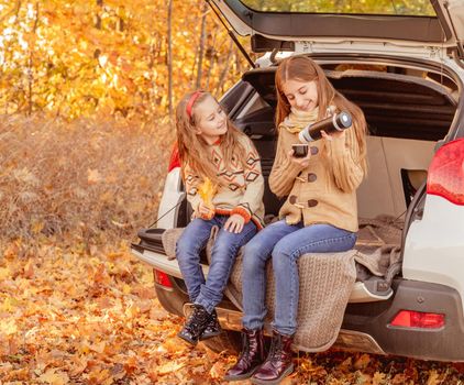 Pretty sisters sharing tea from thermos sitting in car trunk in autumn park