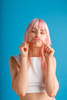 Playful young woman with natural long pink dyed hair holding a strand of it as a moustache, posing isolated over blue studio background. Beauty, hair care concept