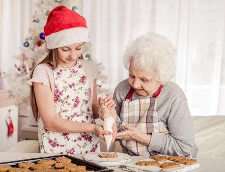 Grandmother with granddaughter in santa hat decorate cookies with cream together