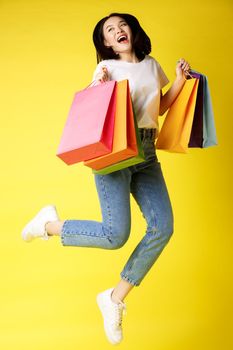 Full size shot of beautiful asian woman jumping with shopping bags and smiling happy, yellow background.