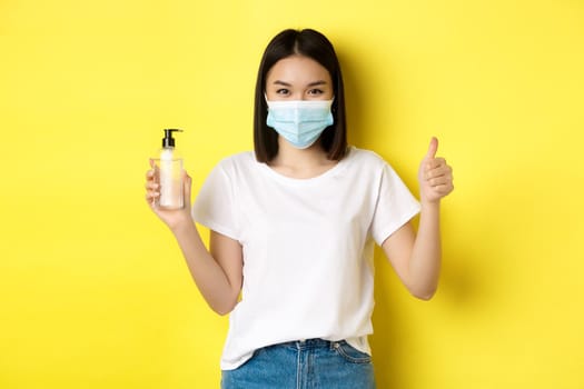 Covid, health care and pandemic concept. Asian girl in face mask from coronavirus, showing thumb up and good hand sanitizer, standing over yellow background.