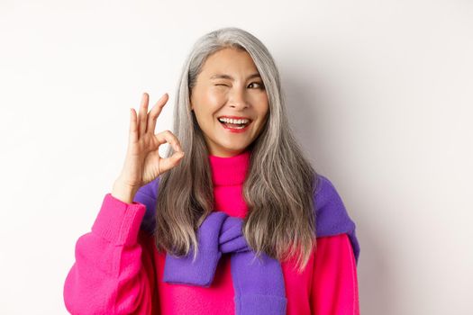 Close-up of cheerful asian grandmother in fashionable pink sweater, winking and showing okay sign, recommending promotion, standing over white background.