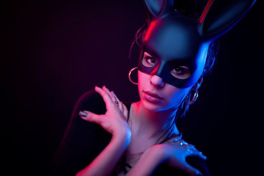 the beautiful girl in a bodysuit poses in a photo Studio on a dark background in neon light. On the face of a rabbit mask