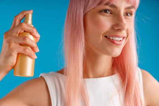 Close up portrait of beautiful young woman with pink hair smiling aside and holding bottle of moisturizing spray, posing isolated over blue studio background. Beauty products, hair care concept