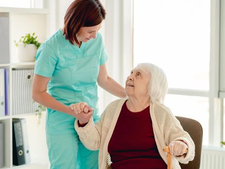 Old lady leaning on nurse hand to stand from chair in nursing home