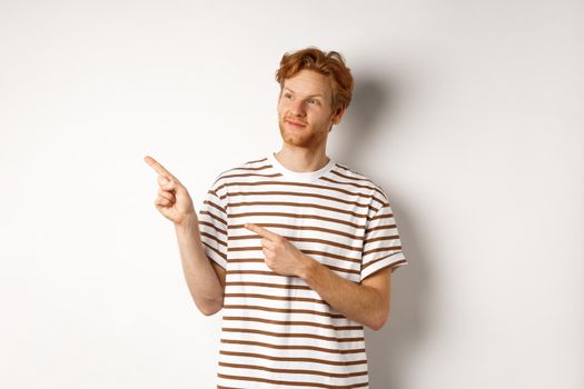 Smiling man with curly red hair, wearing striped t-shirt, smiling and pointing fingers left, demonstrate banner, standing over white background.